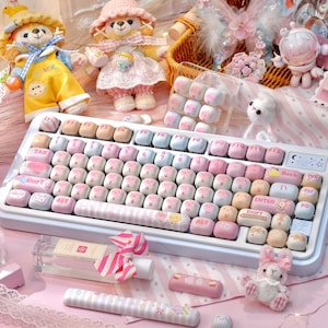 May include: A pastel-colored mechanical keyboard with custom keycaps in shades of pink, blue, and beige. The keyboard is surrounded by plush teddy bears, decorative items, and a perfume bottle. The keycaps have various symbols and text.