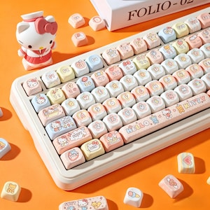 Pode incluir: Um teclado personalizado com teclas em tons pastel com designs Hello Kitty e outros gráficos fofos. O teclado tem uma base branca e rosa, com teclas adicionais espalhadas. Uma estatueta Hello Kitty também está presente.