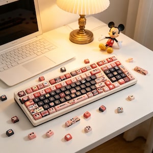 Può includere: Una tastiera meccanica personalizzata con telaio bianco e tasti neri, con tasti rosa e rossi con disegni di Topolino. Tasti aggiuntivi sono sparsi intorno alla tastiera. Sono visibili anche una statuina di Topolino e un laptop.
