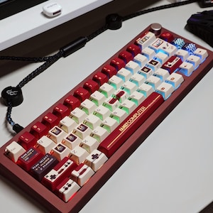 May include: A mechanical keyboard with a maroon frame and a custom keycap set featuring white and maroon keys. The spacebar displays the text FAMILY COMPUTER Nintendo. Several keys include retro gaming graphics, icons, and game controller motifs.