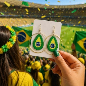 Op de afbeelding: Gouden oorringen met groene en gele avocado-vormige hangers. Elke hanger heeft een ontwerp van de Braziliaanse vlag. De oorbellen worden voor een menigte gehouden, met Braziliaanse vlaggen op de achtergrond.