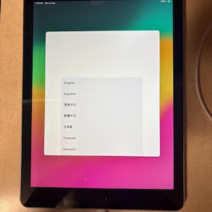 Puede incluir: Una tableta negra que muestra una pantalla de configuración con opciones de idioma: English, Español, 简体中文, 繁體中文, 日本語, Français y Deutsch. La pantalla tiene un fondo degradado de color rosa, verde y amarillo.