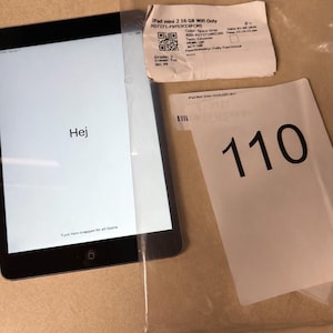 May include: An iPad Mini 2 with 16 GB storage, protected by a screen cover. The display shows "Hej". Two white tags are present: one with the number "110", the other with product information. The tablet is black.