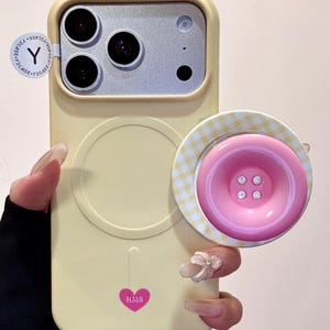 May include: A light yellow phone case with a pink button-shaped phone grip. The phone case has a heart-shaped logo with the letters "HMS". The phone grip has a gingham pattern and four small buttons.