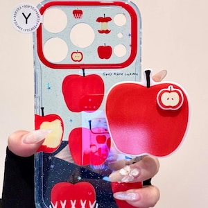 Puede incluir: Una funda de teléfono transparente con un marco de cámara rojo y varias ilustraciones de manzanas rojas, incluyendo manzanas enteras, cortadas y envueltas. El texto "Good Apple Luck day" está impreso y tiene un agarre rojo en forma de manzana.