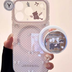 May include: A clear phone case with a pink polka dot pattern and white bows. The case features a teddy bear and bow design near the camera. A matching phone grip with a cat graphic is attached. Text on the grip reads "Angel Feline Form".
