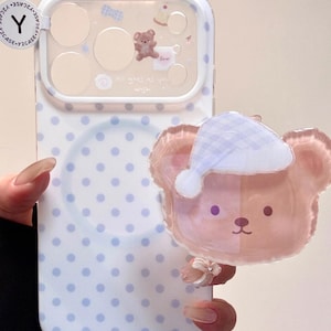 May include: A white and light blue phone case with a polka dot pattern. The case features a clear section with a small bear illustration and the text "All goes as you wish". A pink bear with a white hat is attached.