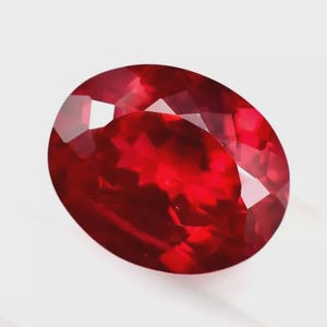 以下が含まれることがあります： ファセットカットされた楕円形のルビーの宝石。宝石は深く鮮やかな赤色で、光を反射する複数のファセットがあります。宝石は無地の白い背景に配置されています。