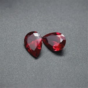 May include: Two deep red, pear-shaped gemstones with multifaceted surfaces. Each stone features a pointed top and a rounded base, reflecting light through its precise geometric cuts to create a brilliant, saturated crimson appearance.
