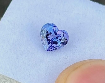 May include: A heart-shaped gemstone in shades of blue and purple. The gem is cut to reflect light, creating a sparkling effect. It sits on a white surface, with a finger partially visible in the image.