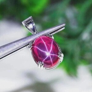 May include: A pendant featuring an oval-shaped, deep magenta star ruby gemstone with a distinct six-rayed star pattern. The cabochon stone is set in a silver-toned metal prong setting with a triangular bail at the top for a chain attachment.