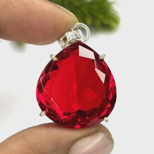 May include: A faceted, teardrop-shaped, red gemstone pendant with a silver bail. The gemstone is held in place by four small gold-colored prongs. The pendant is held between two fingers.