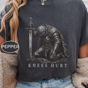 Może przedstawiać: Ciemnoszary t-shirt z krótkim rękawem z grafiką przedstawiającą rycerza w pełnej zbroi, klęczącego i trzymającego miecz. Pod ilustracją znajduje się napis "KNEES HURT" wykonany białymi wielkimi literami. Mała owalna etykieta zawiera napis "PEPPER".