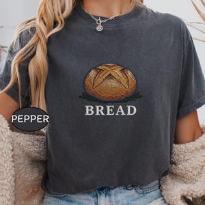 Könnte beinhalten: Ein anthrazitfarbenes Kurzarm-T-Shirt mit einer Stickerei eines runden Brotlaibs, der oben kreuzförmig eingeschnitten ist. Das Wort BREAD ist in weißen Großbuchstaben unter dem Brot eingestickt. Ein kleines schwarzes ovales Etikett trägt die Aufschrift PEPPER.
