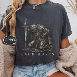 Może przedstawiać: Ciemnoszary t-shirt z krótkim rękawem z grafiką przedstawiającą klęczącego rycerza w zbroi trzymającego miecz. Pod rycerzem znajduje się napis BACK HURTS białymi wielkimi literami. Koszulka ma okrągły dekolt i wykończenie w stylu vintage.