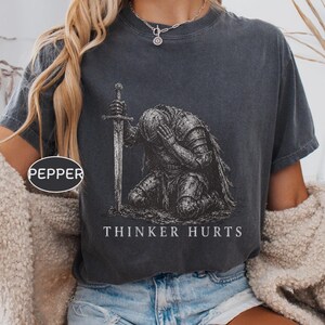 Może przedstawiać: Ciemnoszary t-shirt z krótkim rękawem z grafiką rycerza w zbroi klęczącego z mieczem. Tekst "PEPPER" znajduje się w małym owalu, a słowa "THINKER HURTS" są nadrukowane białymi wielkimi literami poniżej postaci rycerza.