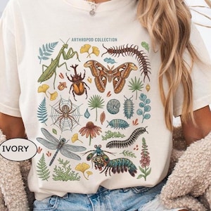 May include: An ivory t-shirt featuring a colourful graphic print titled ARTHROPOD COLLECTION. The design displays various insects, arachnids, and crustaceans including a moth, dragonfly, spider, beetle, and centipede, surrounded by botanical elements.
