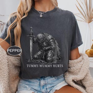 Może przedstawiać: Ciemnoszary t-shirt z krótkim rękawem z grafiką rycerza w zbroi klęczącego i trzymającego miecz. Poniżej rycerza znajduje się napis "TUMMY-WUMMY HURTS" wykonany szeryfową czcionką. Mała owalna etykieta po lewej stronie zawiera słowo "PEPPER".
