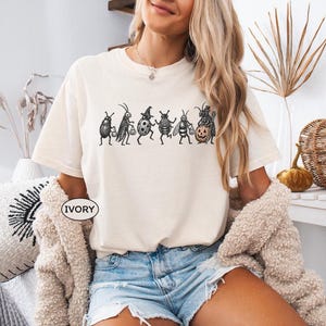 Könnte beinhalten: Ein elfenbeinfarbenes Kurzarm-T-Shirt mit einer schwarzen Grafik, die sechs Insekten in Halloween-Kostümen zeigt, darunter ein Marienkäfer mit Hexenhut und ein Käfer mit einem Kürbis. Das T-Shirt hat einen Rundhalsausschnitt und eine lockere Passform.