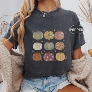 May include: A charcoal grey short-sleeve t-shirt featuring a graphic print of nine pumpkins in various colours and patterns, including floral, celestial, and geometric designs. The shirt has the word PEPPER printed in a white circle on the upper right.