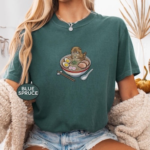 May include: A blue spruce colored short-sleeve t-shirt featuring a graphic illustration of a brown cat eating a bowl of ramen noodles with chopsticks and a spoon. The bowl contains eggs, meat, and pink fish cakes. The text BLUE SPRUCE is visible.