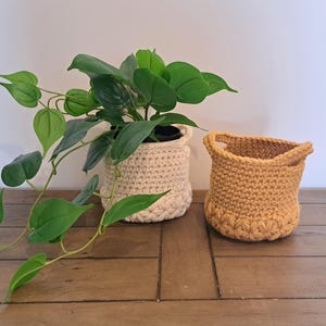 May include: Two handmade crochet plant pots. One is cream and holds a green plant with large leaves. The other is mustard yellow and empty. Both pots are on a wooden surface, creating a rustic, natural aesthetic.