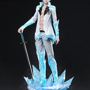 Puede incluir: Figura detallada de un personaje con cabello blanco, vistiendo un abrigo blanco con detalles azules como hielo y pantalones grises. La figura sostiene una espada y se encuentra sobre una base de cristales de hielo. El logo de Tanuki Figures es visible.