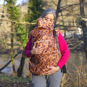 Peut inclure: Un porte-bébé à imprimé léopard marron et noir avec une capuche. Le porte-bébé a une bordure blanche et est porté par une personne portant un pull rose et un pantalon gris. L'arrière-plan présente des arbres et une rivière.