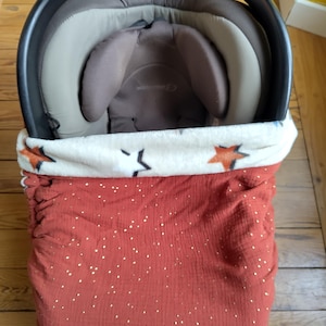 Puede incluir: Un asiento de coche para bebé con una funda de color óxido con un estampado de estrellas blancas y naranjas. El interior del asiento del coche es beige y marrón. El asiento del coche está sobre un suelo de madera.