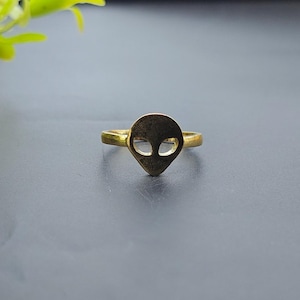 May include: A gold-toned metal ring featuring a flat, stylized alien head motif as the centerpiece. The alien head design includes two large, hollowed-out, almond-shaped eye cutouts. The band is thin and circular, connecting to the base of the head.