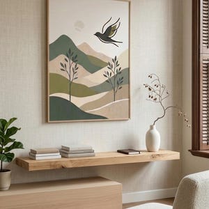 A framed print of a minimalist landscape with green and beige hills, trees, a sun, and a black bird, hangs above a floating wooden shelf in a room.