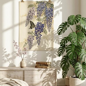 K&ouml;nnte beinhalten: Ein gerahmter Kunstdruck mit einer Aquarellillustration von Blauregenbl&uuml;ten und einem Schmetterling. Das Kunstwerk ist in Lila- und Gr&uuml;nt&ouml;nen gehalten, mit einem beigen Hintergrund. Der Druck h&auml;ngt &uuml;ber einem Holzschrank, mit einer Topfpflanze und B&uuml;chern.