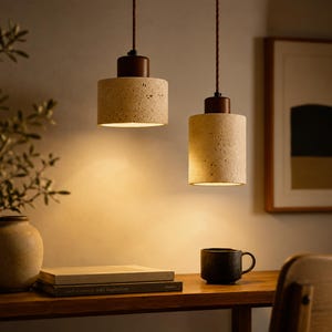 Puede incluir: Dos lámparas colgantes cilíndricas con pantallas beige texturizadas y tapas de madera marrón oscuro cuelgan de cables marrones. Las luces iluminan un escritorio de madera con un jarrón, libros y una taza negra. El fondo es una pared neutra con una obra de arte enmarcada.