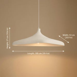 Op de afbeelding: Een witte hanglamp met een brede, ondiepe kap. De lamp is 100 cm lang, 53 cm breed en 27 cm hoog. De armatuur hangt aan een enkele draad en is ontworpen om omgevingslicht te geven.