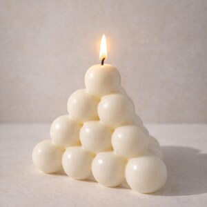 Puede incluir: Una vela blanca con forma de burbujas con una mecha encendida. La vela tiene forma de pirámide, con múltiples esferas apiladas. La llama es amarilla y naranja.