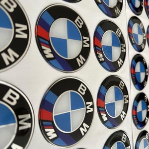 Könnte beinhalten: Mehrere BMW-Emblem-Aufkleber. Jeder Aufkleber hat einen schwarzen Rand, das BMW-Logo in Blau und Weiß und die Buchstaben "BMW" in Weiß. Einige Aufkleber haben rote und blaue Streifen.