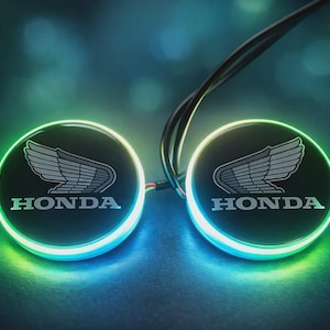 May include: Two illuminated Honda emblems with a glowing green and blue edge. The Honda logo, featuring a stylized wing design, is centered on a black background. The emblems are connected by wires, suggesting an electrical component or accessory.