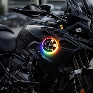 Puede incluir: Una motocicleta negra con un emblema iluminado multicolor en la parte delantera. El emblema es un círculo con una cruz y las letras "NI" y "M" en su interior. La motocicleta tiene parabrisas y faros.