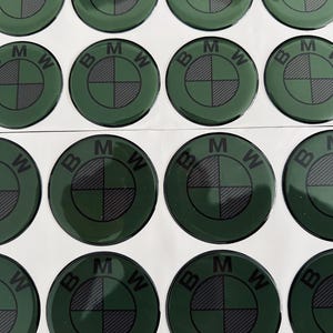 Puede incluir: Una colección de pegatinas con el emblema de BMW en verde y negro. Cada pegatina circular presenta el logotipo de BMW, con las letras "BMW" y un patrón de cuadros en el centro. Las pegatinas están dispuestas en filas y columnas.