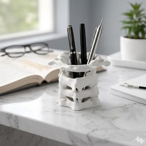Peut inclure: Un porte-stylos blanc en forme de colonne vertébrale contenant trois stylos. Le support est en céramique et repose sur une surface en marbre. Les stylos sont noirs et argentés. Un livre et des lunettes sont en arrière-plan.