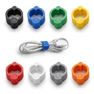 Puede incluir: Una colección de organizadores de cables coloridos en negro, azul, verde, amarillo, rojo, blanco, gris y naranja. Un organizador azul asegura un cable blanco con un conector USB. Los organizadores son circulares con un cierre y un botón central.
