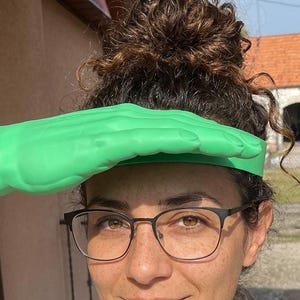 Può includere: Una persona indossa una visiera verde a forma di mano. La persona indossa occhiali con montatura nera e una camicia di colore chiaro. Lo sfondo include una struttura in legno e un edificio.
