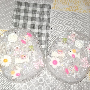 May include: Two clear, oval resin coasters adorned with miniature charms. The charms feature teddy bears, flowers, cherries, bows, and other cute decorations in pink, white, and yellow. The coasters are displayed on a patterned background.