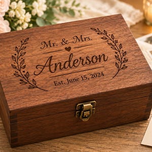 Peut inclure: Une boîte à souvenirs rectangulaire en bois brun foncé avec un fermoir en métal. Le couvercle est gravé au laser avec le texte Mr. & Mrs. Anderson, une icône de cœur, une couronne florale et la date Est. June 15, 2024, centrée sur le dessus.