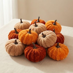 May include: A collection of eleven plush pumpkins in various shades of orange, rust, and beige. Each pumpkin features a soft, textured fabric surface and a dark brown, smooth, rigid stem protruding from the centre of its rounded, ribbed body.
