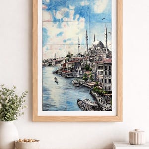 Op de afbeelding: Ingelijst kunstwerk dat een stadsgezicht aan het water toont met een moskee en minaretten. De schildering staat op tegels, omlijst in licht hout. De lucht is blauw met wolken en er varen boten op het water.