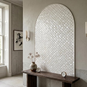 May include: An arched wall feature covered in small, iridescent white oval tiles arranged in a repeating pattern. The structure is framed by a thin border and sits above a dark wooden console table holding a ceramic vase and two small round clocks.
