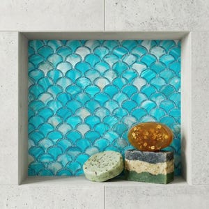 Puede incluir: Tres pastillas de jabón colocadas en un nicho de pared con un patrón de azulejos de escamas de pez en color turquesa brillante. Un jabón es ovalado y de color ámbar con inclusiones, otro es redondo y moteado, y otro es rectangular y estratificado.