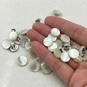 May include: A collection of round, white, iridescent mother-of-pearl buttons with a smooth, polished surface. Each button features a central shank on the back for attachment. The buttons have a subtle, pearlescent sheen and a uniform circular shape.