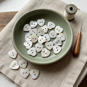 May include: A collection of small, heart-shaped white mother-of-pearl buttons, each featuring two central holes. The buttons are arranged inside a round, sage green ceramic bowl, with several additional buttons scattered outside the bowl.